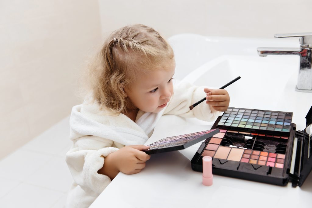 Toddler girl doing herself makeup with mama's cosmetics pretending grownup. Applying eyeshadows. Child in the bathrobe at home